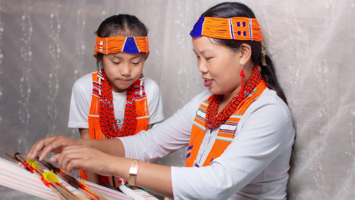 A mother and her daughter wearing traditional orange beaded Konyak Naga attire sit together indoors, working on a handloom weaving frame. Both wear matching beaded headbands and necklaces, while the woman carefully demonstrates the weaving process to the child against a soft neutral background.